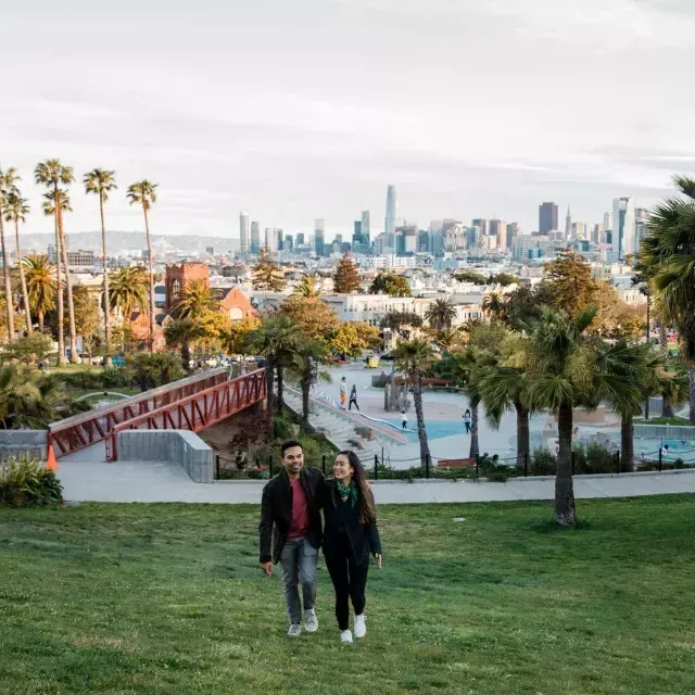 一对夫妇走向镜头，Dolores Park和威尼斯人官网平台app的天际线在他们身后.